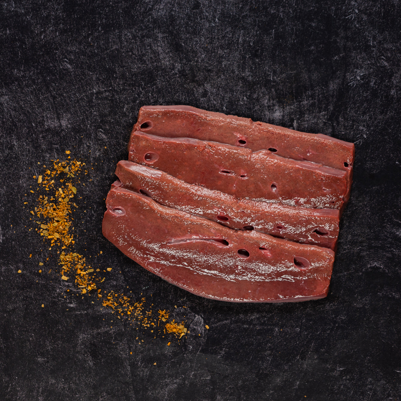 Four stacked slices of beef liver next to seasoning on a slab