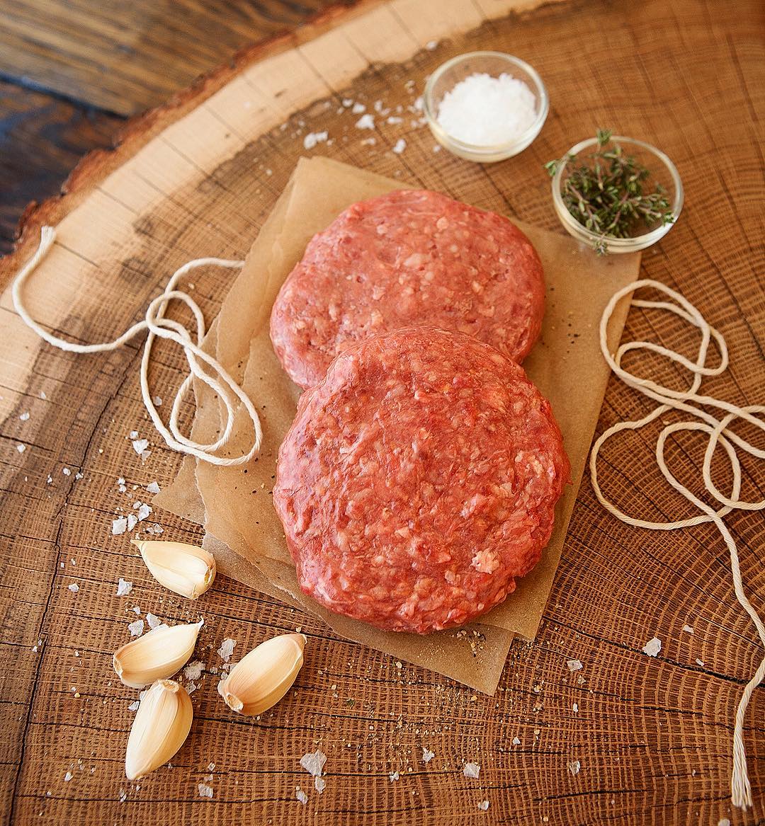85/15 Ground Beef in 2 patties partially laying on top of each other on top of kraft parchment paper. Garlic cloves, twine, and salt flakes and thyme in small glass containers are outlining the patties.
