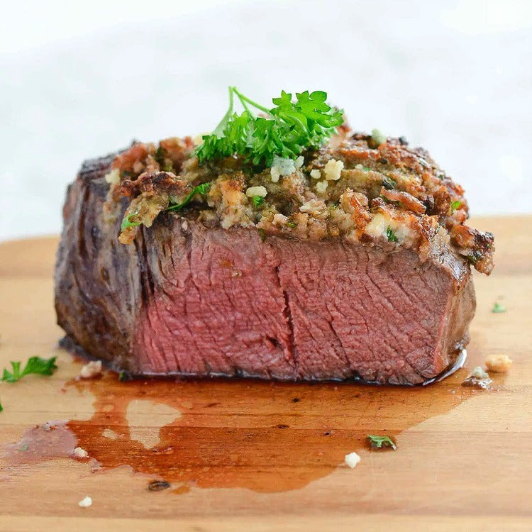 Bison Filet Mignon with sautéed garlic  on top garnished with a piece of parsley, sitting on a wood cutting board and cut showing a perfect Medium temperature with a little juice that has leaked out 